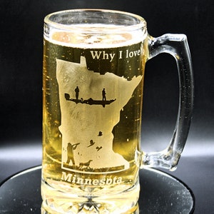May include: A clear glass beer stein with a handle. The stein is filled with a golden-colored beer. The stein is etched with an outline of the state of Minnesota, with the words "Why I love Minnesota" and images of people fishing and hunting.