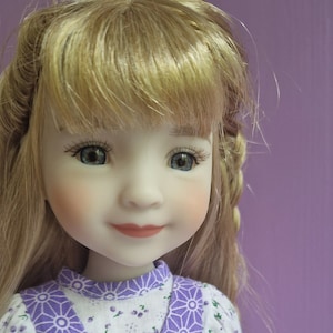 Op de afbeelding: Een pop met blond haar die een paars-witte jurk draagt met een witte strik en een zilveren vlinderbedel.