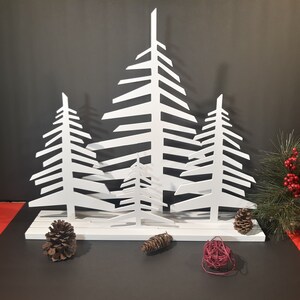 May include: White metal Christmas tree silhouette decoration with four trees of varying sizes. The trees are cut out and stand on a white base.