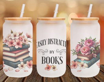 Könnte beinhalten: Drei Glasbecher mit Bambusdeckeln und Strohhalmen. Jeder Becher hat ein anderes Design: einer mit einem Stapel Bücher und rosa Blumen, einer mit dem Text "Easily Distracted by Books" und einer mit einem Strauß rosa Blumen und einem Stapel Bücher.