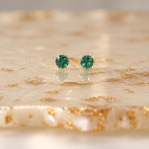 May include: A pair of stud earrings featuring round, faceted teal-green gemstones set in gold-toned metal prongs. The earrings are positioned side-by-side, showcasing the brilliant cut of the stones and the metallic luster of the settings.