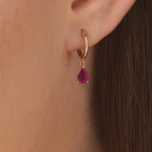 Puede incluir: Un pendiente de aro dorado con un pequeño dije de piedra preciosa en forma de lágrima de color rosa intenso suspendido en la parte inferior. El pendiente está sujeto al lóbulo de la oreja, mostrando un acabado metálico pulido y una piedra facetada.