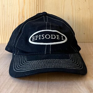 Peut inclure: Casquette de baseball noire avec des coutures blanches et un patch ovale blanc qui dit "EPISODE I" en noir.