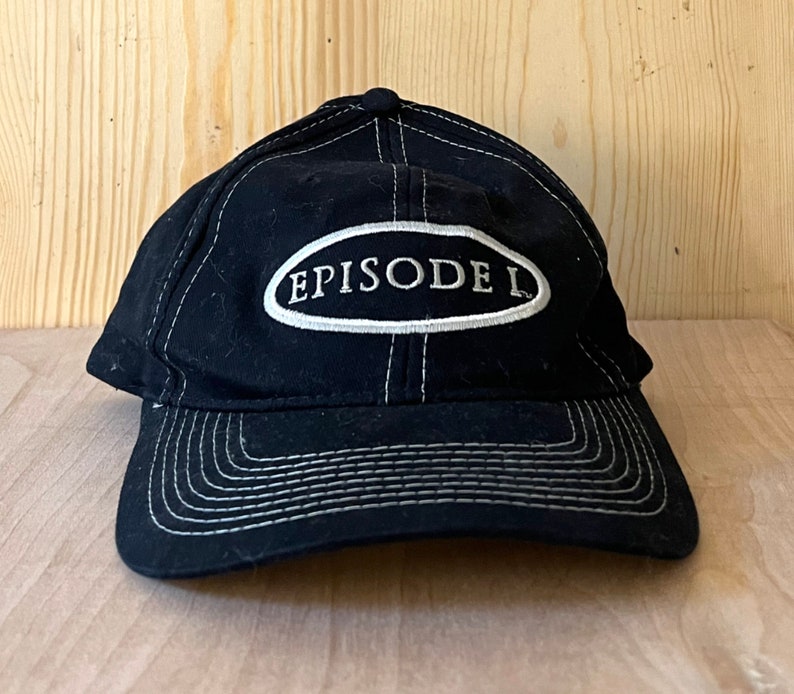 Peut inclure: Casquette de baseball noire avec des coutures blanches et un patch ovale blanc qui dit "EPISODE I" en noir.
