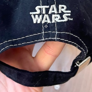 Peut inclure: Casquette de baseball noire avec un logo "STAR WARS" brod&eacute; en blanc sur le devant. La casquette a un d&eacute;tail de couture blanche autour du bord et une fermeture &agrave; boucle en m&eacute;tal argent&eacute;.