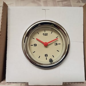 Può includere: Un orologio rotondo con quadrante color crema, numeri neri e lancette rosse. L'orologio è inserito in una lunetta cromata ed è confezionato in una scatola di cartone con inserti in schiuma bianca. L'orologio è probabilmente per un veicolo.