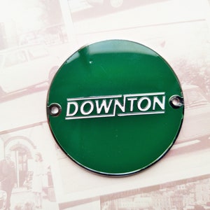 Puede incluir: Un emblema redondo de metal verde con una inscripción blanca que dice "DOWNTON".