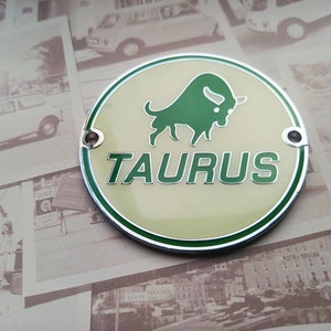 Puede incluir: Un emblema redondo de metal de color crema con un borde verde. El emblema presenta una silueta de toro verde y la palabra "TAURUS" en letras verdes.