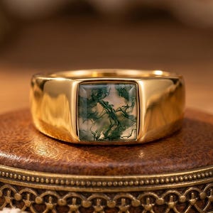 Peut inclure: Une bague en or ornée d'une pierre d'agate mousse de forme carrée avec des inclusions vertes rappelant des fougères. L'anneau est lisse, poli et en or massif, présentant un design épuré qui met en valeur les motifs naturels de la pierre.