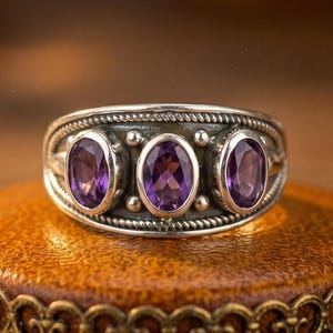 May include: A silver ring featuring three oval-cut purple amethyst gemstones set in a row. The band has a textured, rope-like border design along the top and bottom edges, with small metal bead accents placed between each of the three stone settings.
