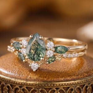 Peut inclure: Une bague en or avec une pierre centrale en agate mousse en forme de poire aux inclusions vert foncé, entourée de petites pierres vertes taille marquise et de pierres blanches rondes disposées en grappe sur un anneau fin en or.