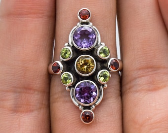 Op de afbeelding: Een zilveren ring met een geometrisch ontwerp en diverse edelstenen. De ring heeft een centrale cluster van paarse, gele, groene en rode stenen, waardoor een levendig sieraad ontstaat. De ring wordt door een hand vastgehouden.
