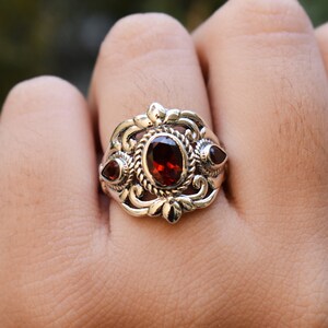 Puede incluir: Un anillo de plata con una piedra granate de color rojo oscuro de corte ovalado en el centro, rodeada por un borde de metal trenzado. Dos piedras granate m&aacute;s peque&ntilde;as en forma de pera est&aacute;n engastadas a los lados con motivos florales.