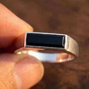Könnte beinhalten: Ein silberner Ring mit einem rechteckigen, horizontal ausgerichteten schwarzen Stein, der in die Ringschiene eingelassen ist. Der Metallring hat eine glatte, polierte Oberfläche und eine flache Oberseite, die den länglichen schwarzen Edelstein sicher hält.