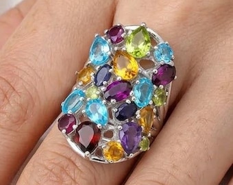 Puede incluir: Un anillo de plata con un conjunto de piedras preciosas multicolores de diversas formas, incluyendo cortes ovalados, de pera y redondos. Las piedras muestran tonos vibrantes de azul, morado, amarillo, verde y rojo, engastadas en un marco de metal plateado.
