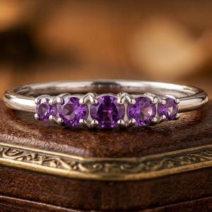 May include: A silver-toned ring featuring five round-cut purple amethyst stones set in a row. The center stone is the largest, with two smaller stones on each side, all held in place by a metal prong setting on a smooth, polished band.