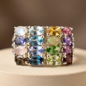 May include: A multi-stone ring featuring four vertical rows of oval-cut gemstones set in a silver-toned metal band. The stones display a variety of colours including blue, green, yellow, pink, purple, and brown, arranged in a grid-like pattern.