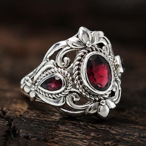May include: A silver ring featuring a central oval-cut deep red garnet gemstone surrounded by a twisted metal border. The band displays intricate openwork scroll patterns and additional smaller pear-shaped garnet stones on the sides.