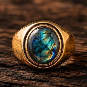 Op de afbeelding: Een goudkleurige ring met een gladde, gepolijste band en een centrale ovaalvormige cabochon edelsteen. De steen vertoont een levendig, iriserend kleurenspel van blauwe en goudgele tinten over het oppervlak, gezet in een eenvoudige metalen zetting.