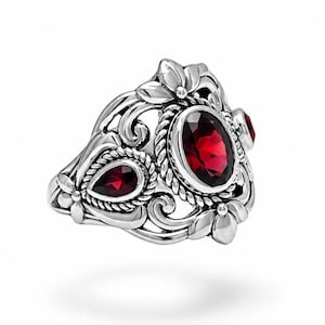 Puede incluir: Un anillo de plata con una piedra roja central de corte ovalado rodeada por un borde de cuerda trenzada. La banda muestra intrincados motivos florales y de volutas, con una piedra roja adicional en forma de pera en el lateral del marco.