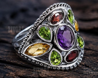 Op de afbeelding: Een zilveren ring met een centrale ovale paarse edelsteen omringd door kleinere markies- en peervormige stenen in groen-, geel- en roodtinten. De metalen band heeft een kralentextuur langs de randen van de decoratieve zetting.