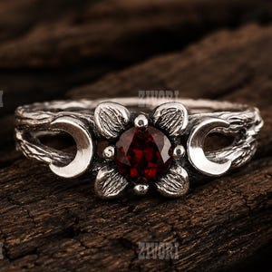 Könnte beinhalten: Ein Silberring mit einem runden, dunkelroten Edelstein in der Mitte, der in ein vierblättriges Blumenmotiv gefasst ist. Die Ringschiene hat eine rindenartige Textur und zeigt zwei Mondsicheln neben der Blume. Der Text ZIVORI ist zu sehen.