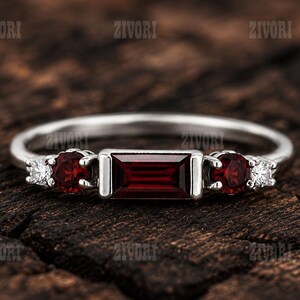 Puede incluir: Un anillo de tono plateado con un granate rojo profundo central de corte baguette rectangular, flanqueado por dos granates rojos redondos y dos pequeñas piedras redondas claras a cada lado. La palabra ZIVORI se repite en el fondo.
