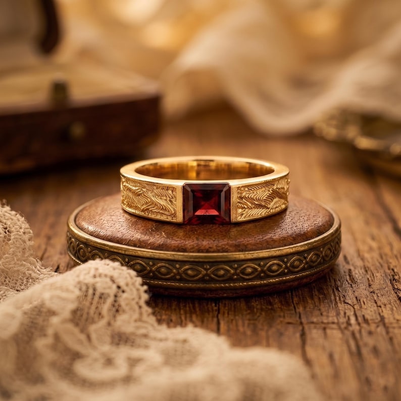 May include: A gold ring featuring a square-cut deep red gemstone set in the center. The band is crafted from yellow gold and displays a detailed, textured pattern of repeating diagonal lines etched across the surface on both sides of the stone.