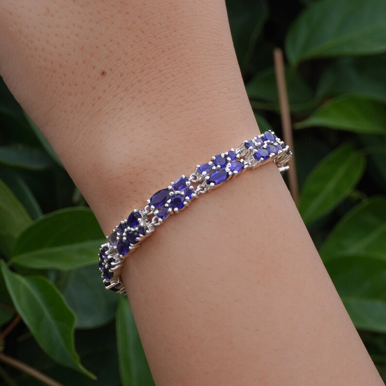May include: A silver bracelet with a double row of oval, deep blue gemstones. The bracelet is worn on a wrist, with a green leafy background. The gemstones are set closely together, creating a continuous band of color.