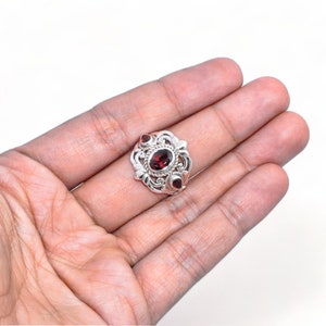 Puede incluir: Un anillo de plata con una piedra preciosa de granate rojo oscuro de corte ovalado en el centro, rodeada de intrincados adornos met&aacute;licos y dos piedras de granate rojas m&aacute;s peque&ntilde;as en forma de l&aacute;grima a los lados del engaste.