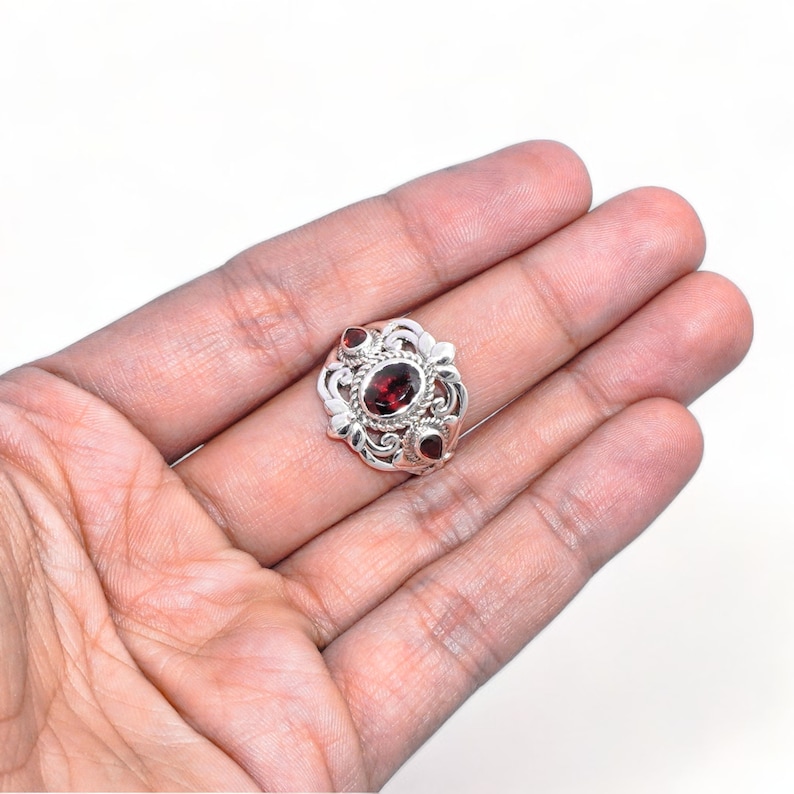 Puede incluir: Un anillo de plata con una piedra preciosa de granate rojo oscuro de corte ovalado en el centro, rodeada de intrincados adornos met&aacute;licos y dos piedras de granate rojas m&aacute;s peque&ntilde;as en forma de l&aacute;grima a los lados del engaste.