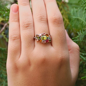 K&ouml;nnte beinhalten: Ein silberner Ring mit einem Cluster aus bunten Edelsteinen, darunter gr&uuml;n, rosa und orange, in einem floralen Design.