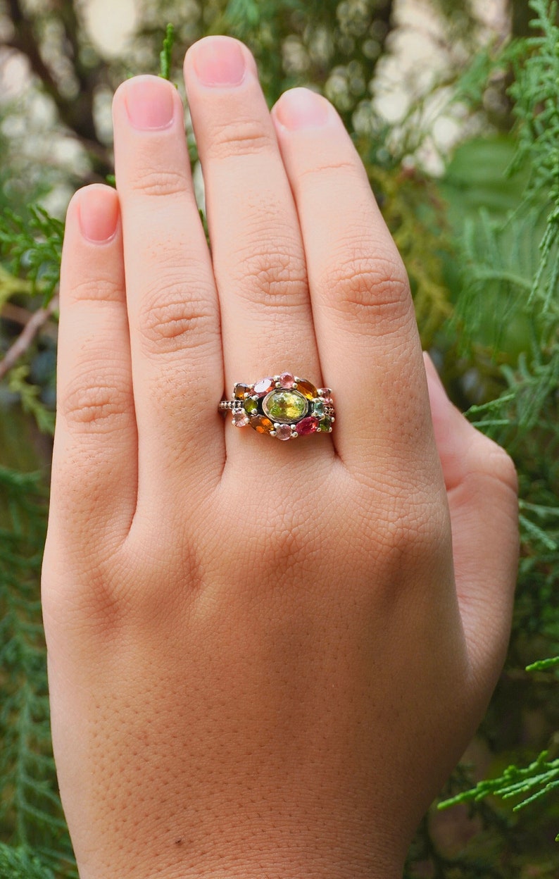 K&ouml;nnte beinhalten: Ein silberner Ring mit einem Cluster aus bunten Edelsteinen, darunter gr&uuml;n, rosa und orange, in einem floralen Design.