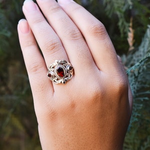 Puede incluir: Un anillo de plata con una gran piedra granate de color rojo oscuro y corte ovalado en el centro, flanqueada por dos piedras granate m&aacute;s peque&ntilde;as en forma de pera. La banda de metal muestra volutas intrincadas y una textura trenzada.