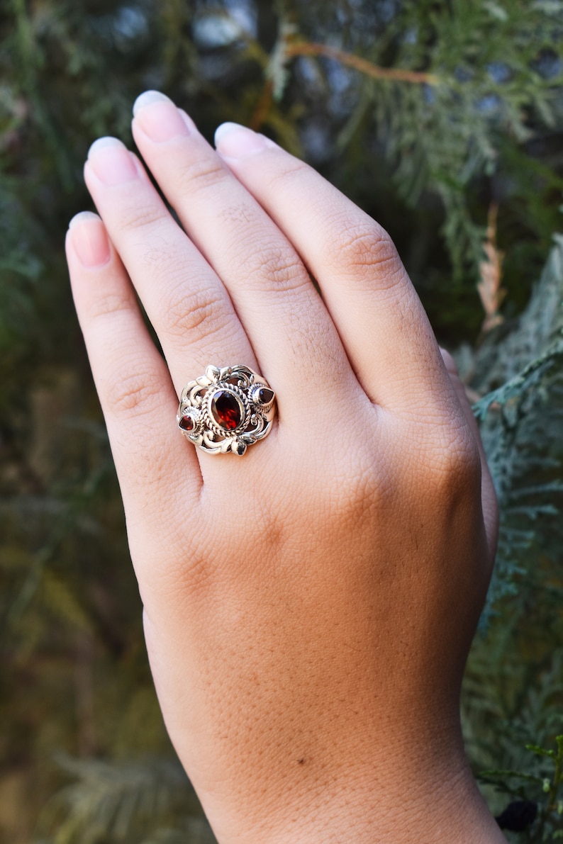 Puede incluir: Un anillo de plata con una gran piedra granate de color rojo oscuro y corte ovalado en el centro, flanqueada por dos piedras granate m&aacute;s peque&ntilde;as en forma de pera. La banda de metal muestra volutas intrincadas y una textura trenzada.