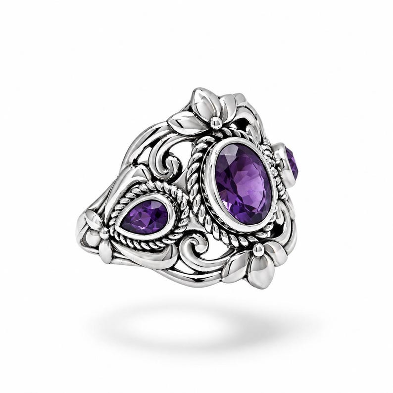 Puede incluir: Un anillo de plata con una gran amatista morada de corte ovalado en el centro, flanqueada por una amatista m&aacute;s peque&ntilde;a en forma de pera. La banda presenta intrincados detalles de metal con volutas, texturas de cuerda y motivos florales.