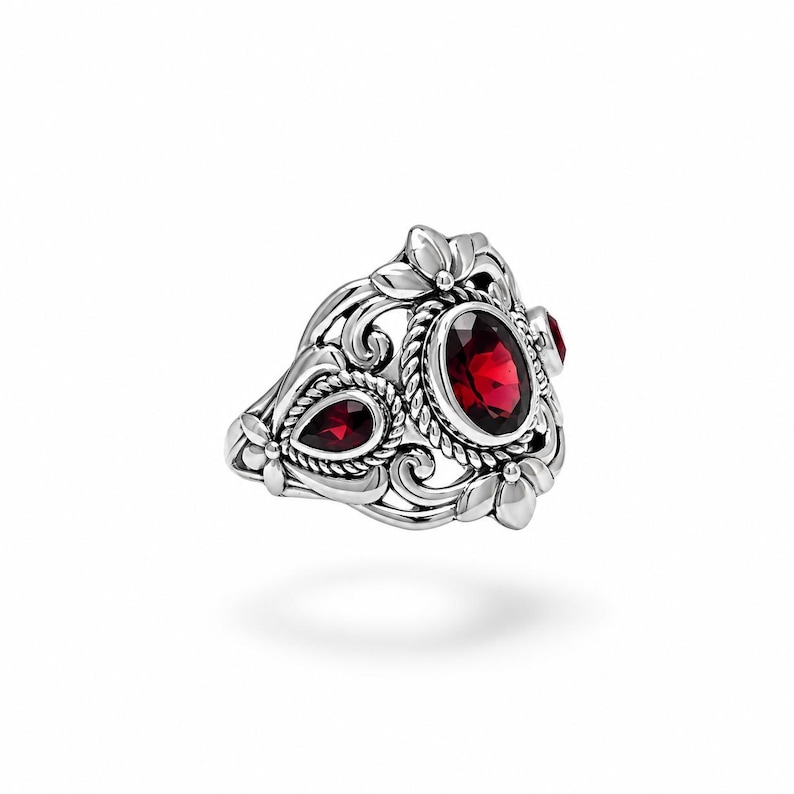 Puede incluir: Un anillo de plata con una piedra roja central de corte ovalado rodeada por un borde de cuerda trenzada. La banda muestra intrincados motivos florales y de volutas, con una piedra roja adicional en forma de pera en el lateral del marco.