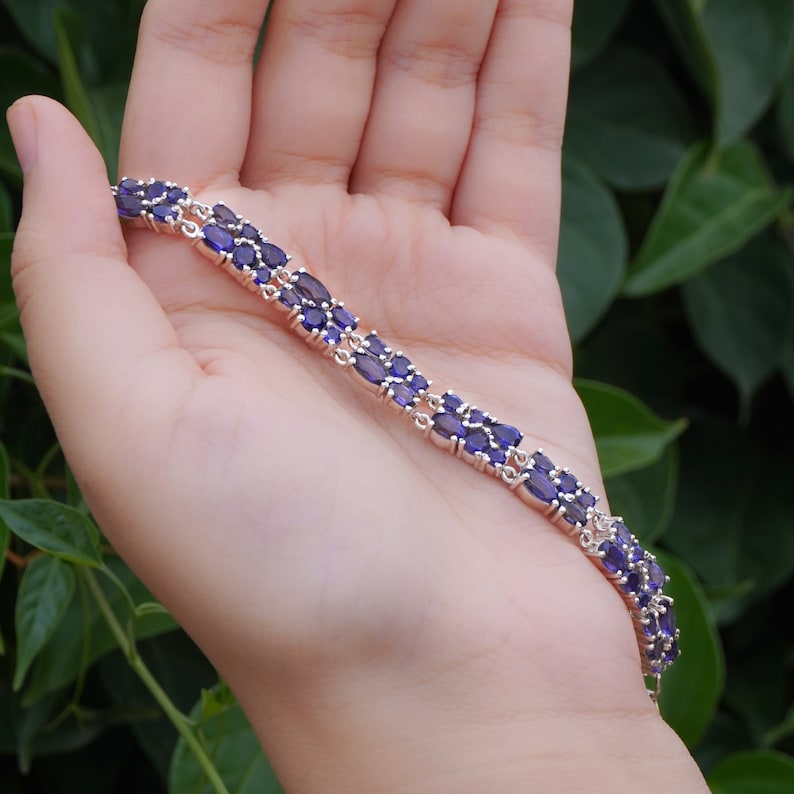 May include: A silver bracelet featuring a double row of oval, deep purple gemstones. The bracelet is held in a hand against a backdrop of green foliage, showcasing the jewelry's design and color.