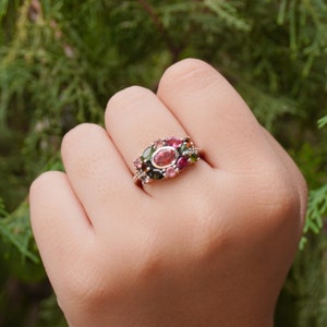 K&ouml;nnte beinhalten: Ein goldener Ring mit einem Cluster aus bunten Edelsteinen, darunter rosa, gr&uuml;n und braun. Der Ring ist mit einem gr&ouml;&szlig;eren ovalen rosafarbenen Edelstein in der Mitte besetzt.