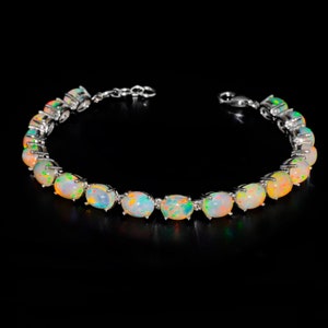 May include: A silver-toned bracelet featuring a series of oval-shaped, iridescent opals set in metal prongs. Each opal displays a play of colour with flashes of orange, green, and blue against a milky white base. The bracelet includes a metal chain link extension and a lobster claw clasp.
