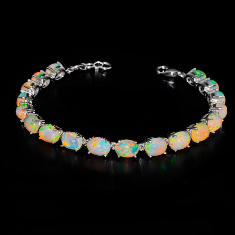 May include: A silver-toned bracelet featuring a series of oval-shaped, iridescent opals set in metal prongs. Each opal displays a play of colour with flashes of orange, green, and blue against a milky white base. The bracelet includes a metal chain link extension and a lobster claw clasp.