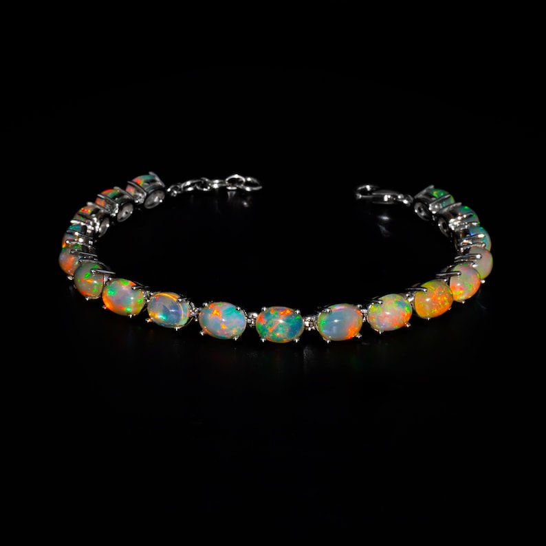 May include: A silver-toned bracelet featuring a series of oval-shaped opals set in metal prongs. Each opal displays a play of colour with flashes of orange, green, and blue against a translucent base. The bracelet includes a metal link chain clasp.