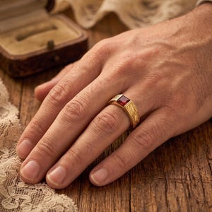 May include: A gold ring featuring a square-cut red gemstone set in the center. The band of the ring has a textured, brushed metal finish. The ring is worn on a finger, showcasing the contrast between the polished red stone and the gold band.
