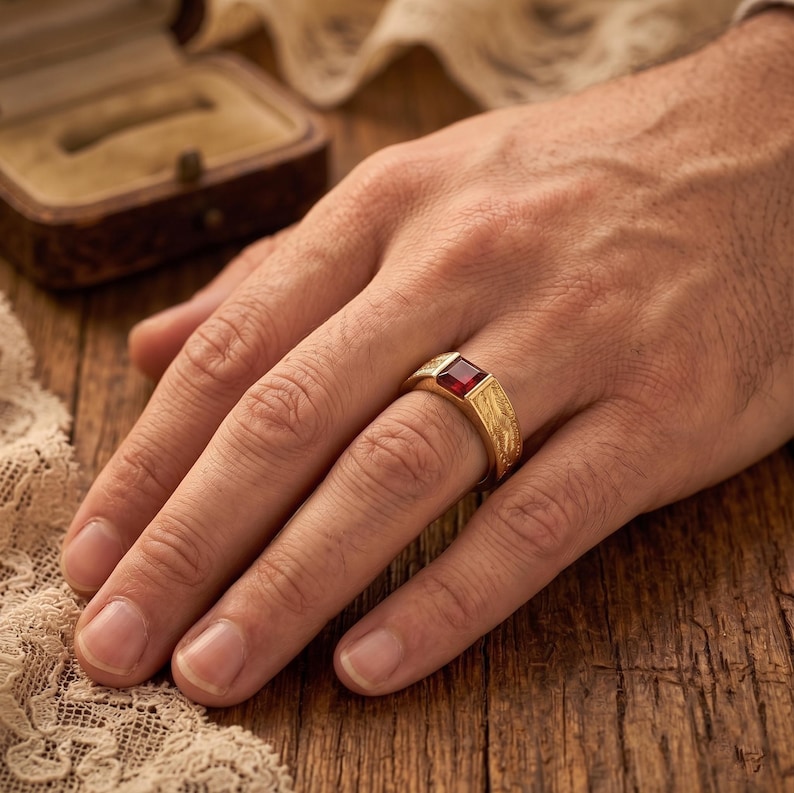 May include: A gold ring featuring a square-cut red gemstone set in the center. The band of the ring has a textured, brushed metal finish. The ring is worn on a finger, showcasing the contrast between the polished red stone and the gold band.