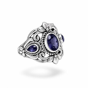 Puede incluir: Un anillo de plata de ley con una gran piedra preciosa de color p&uacute;rpura intenso de corte ovalado rodeada por un borde de cuerda trenzada, flanqueada por piedras p&uacute;rpuras m&aacute;s peque&ntilde;as en forma de pera, sobre un marco de metal con motivos florales.