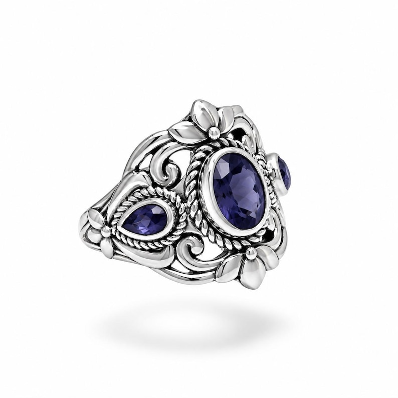 Puede incluir: Un anillo de plata de ley con una gran piedra preciosa de color p&uacute;rpura intenso de corte ovalado rodeada por un borde de cuerda trenzada, flanqueada por piedras p&uacute;rpuras m&aacute;s peque&ntilde;as en forma de pera, sobre un marco de metal con motivos florales.