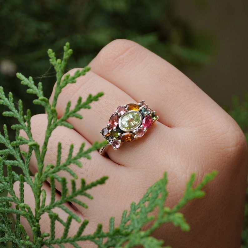 May include: A silver ring featuring a cluster of colourful gemstones, including pink, green, and yellow. The ring is set with a large, oval-shaped green gemstone in the centre.
