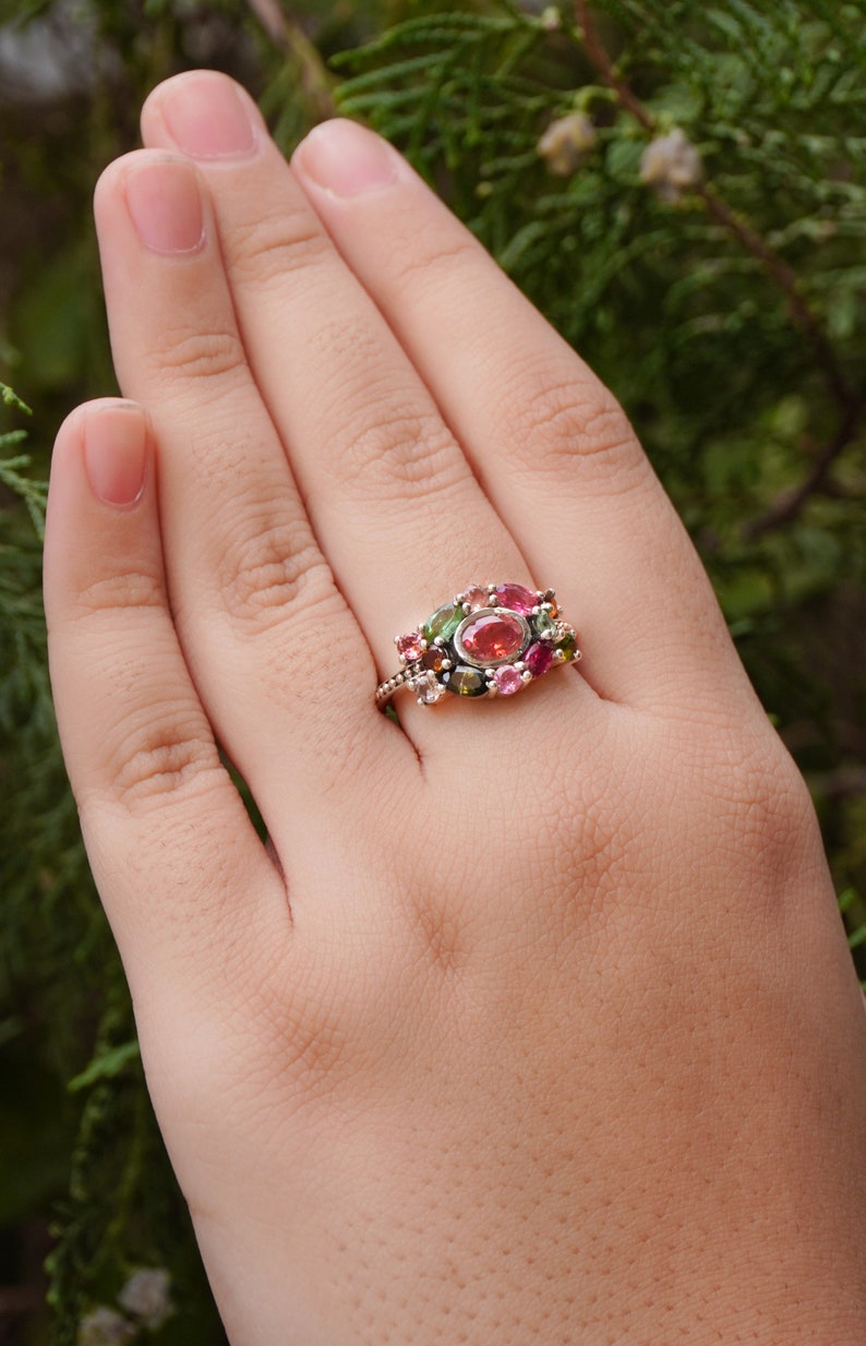 K&ouml;nnte beinhalten: Ein Ring mit einem Cluster aus rosa, gr&uuml;nen und schwarzen Edelsteinen, die in einer silbernen Fassung sitzen.