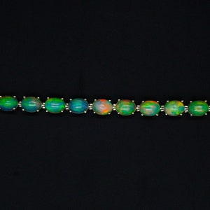May include: A silver-toned bracelet featuring a series of oval-shaped cabochon opals. Each opal displays iridescent flashes of green, blue, and orange. The bracelet is secured with a lobster claw clasp and includes a small circular link extender chain.