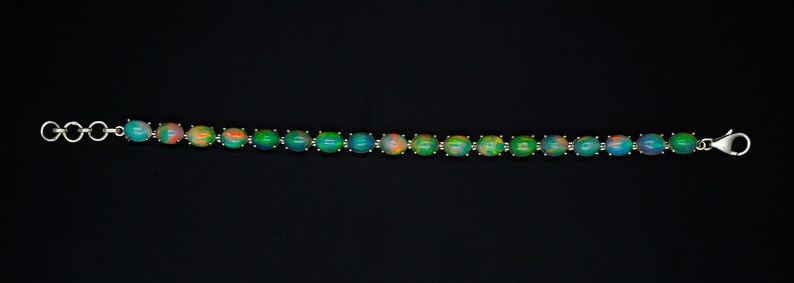 May include: A silver-toned bracelet featuring a series of oval-shaped cabochon opals. Each opal displays iridescent flashes of green, blue, and orange. The bracelet is secured with a lobster claw clasp and includes a small circular link extender chain.